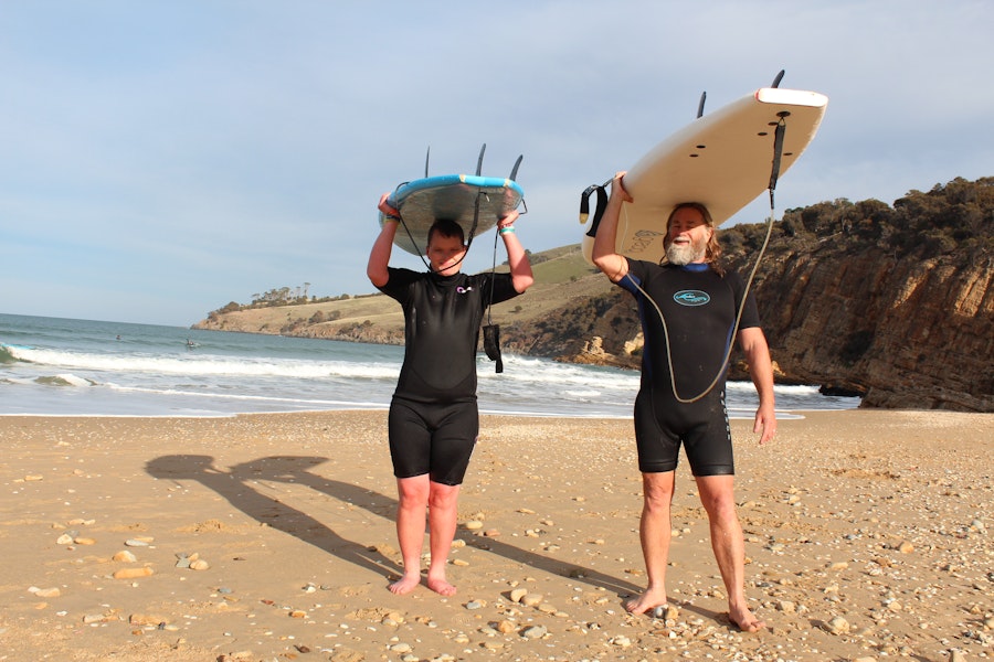 Two persons in wetsuits walking on the beach.  They are holding surfboards that are resting above them on their heads.